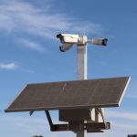 Câmera de vigilância instalada em poste com painel solar, contra o céu azul.