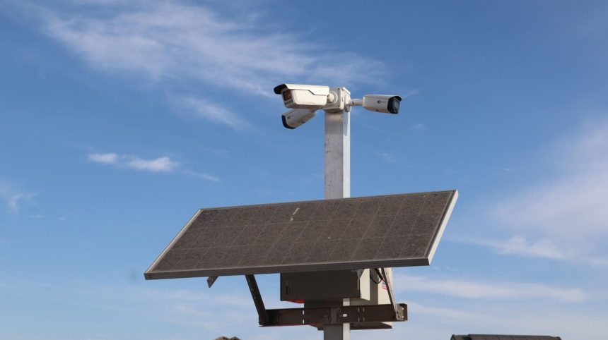 Câmera de vigilância instalada em poste com painel solar, contra o céu azul.